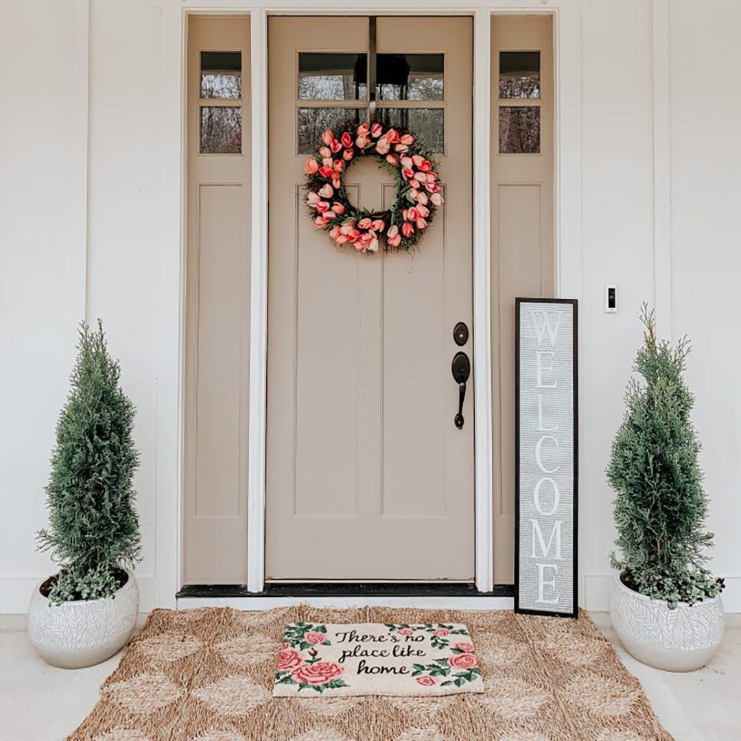 Spring front porch decor with a door mat, stand up welcome sign, a pine tree in ceramic planters on each side of the door and a pink rose wreath on the door.